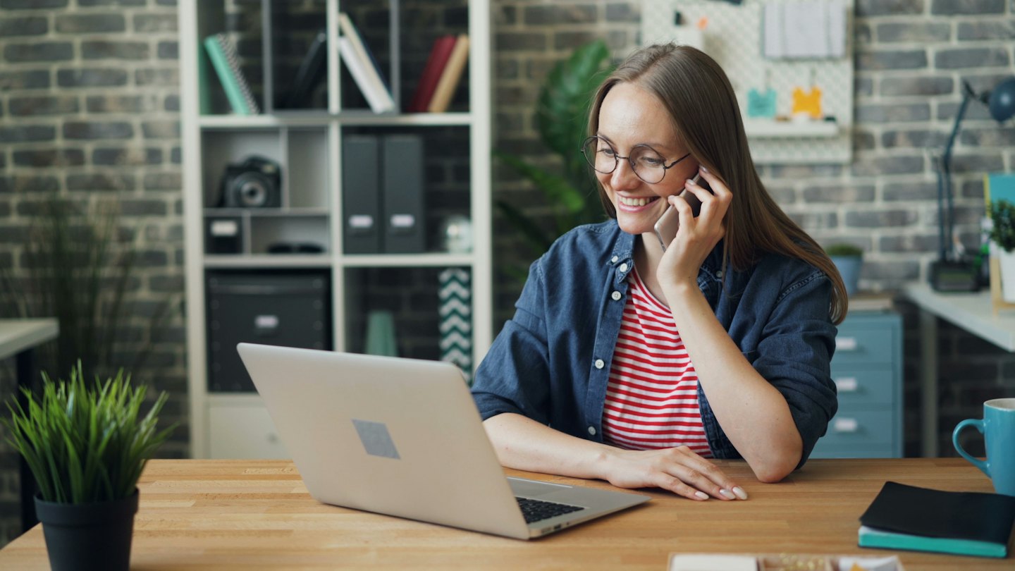 Woman smiles while talking on a smartphone at her desk with a laptop.